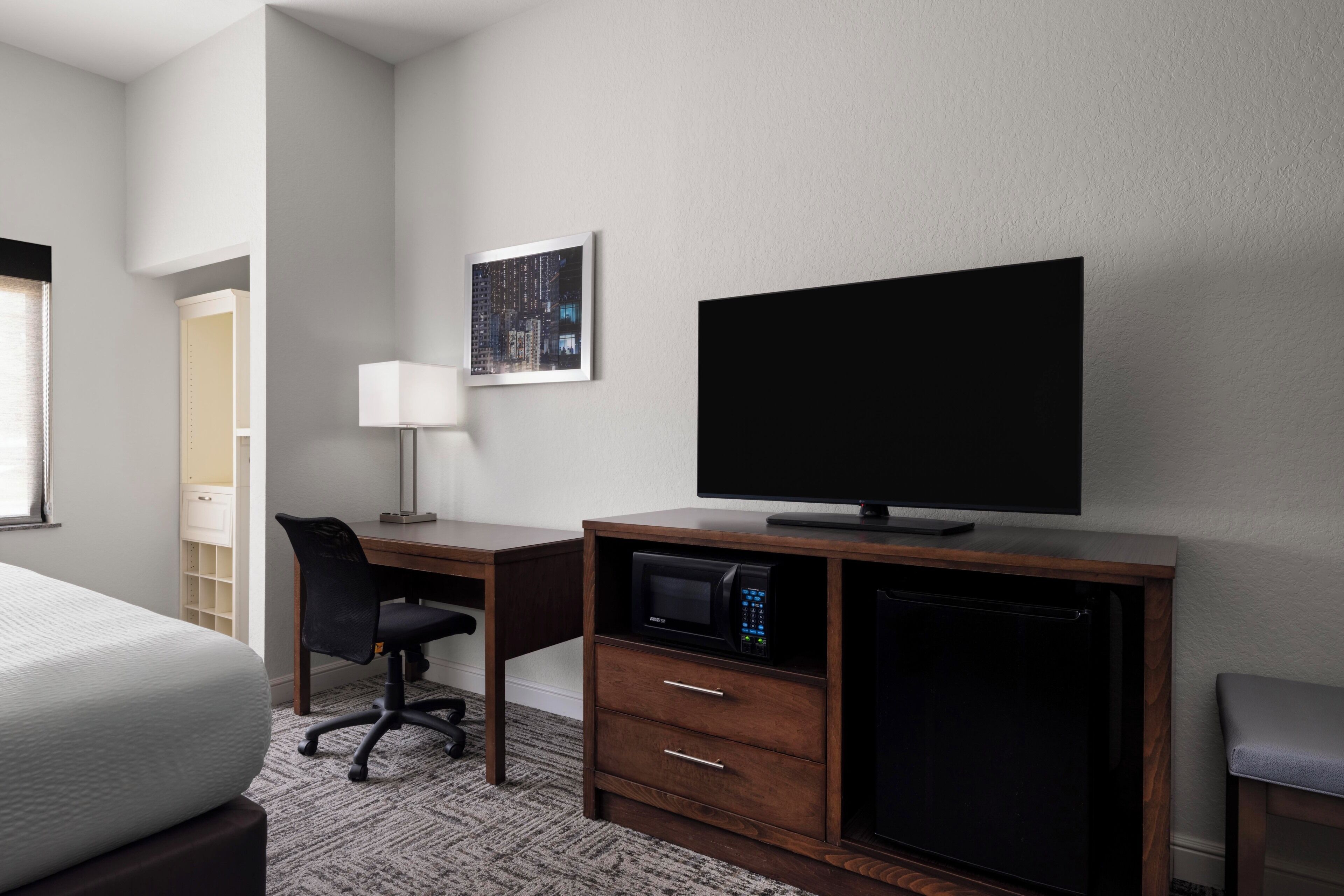 A hotel room with a bed, a desk with a computer, a flat-screen TV, a microwave, and a small refrigerator.