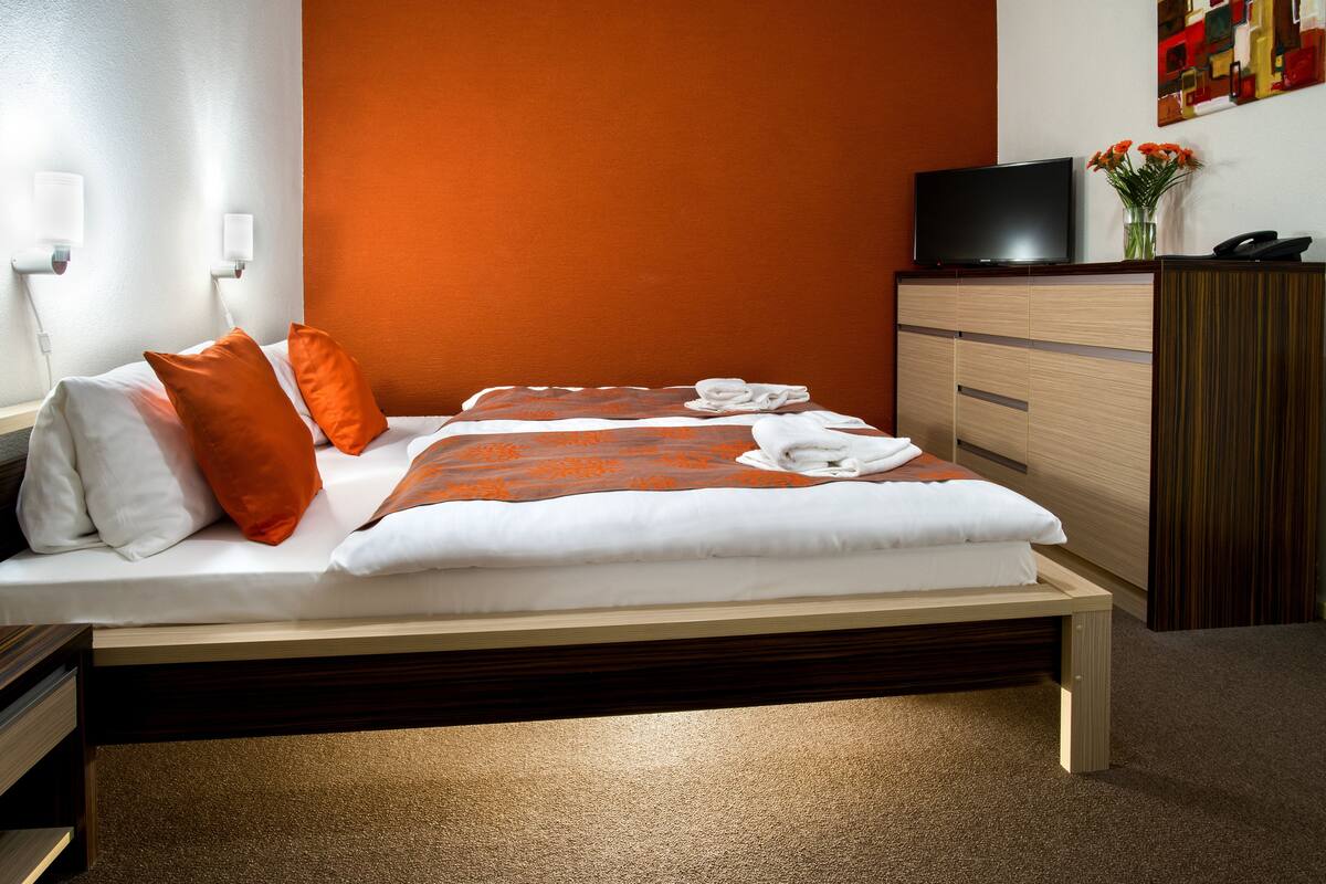 A neatly made bed with white and orange bedding, a television on a wooden cabinet, and a vase with flowers on the wall.