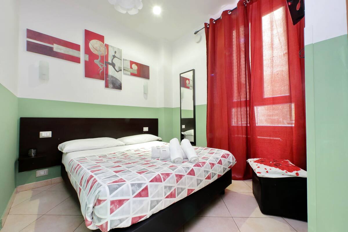 A bedroom with a bed, a bedside table, a nightstand, a red curtain, and a window with a view.