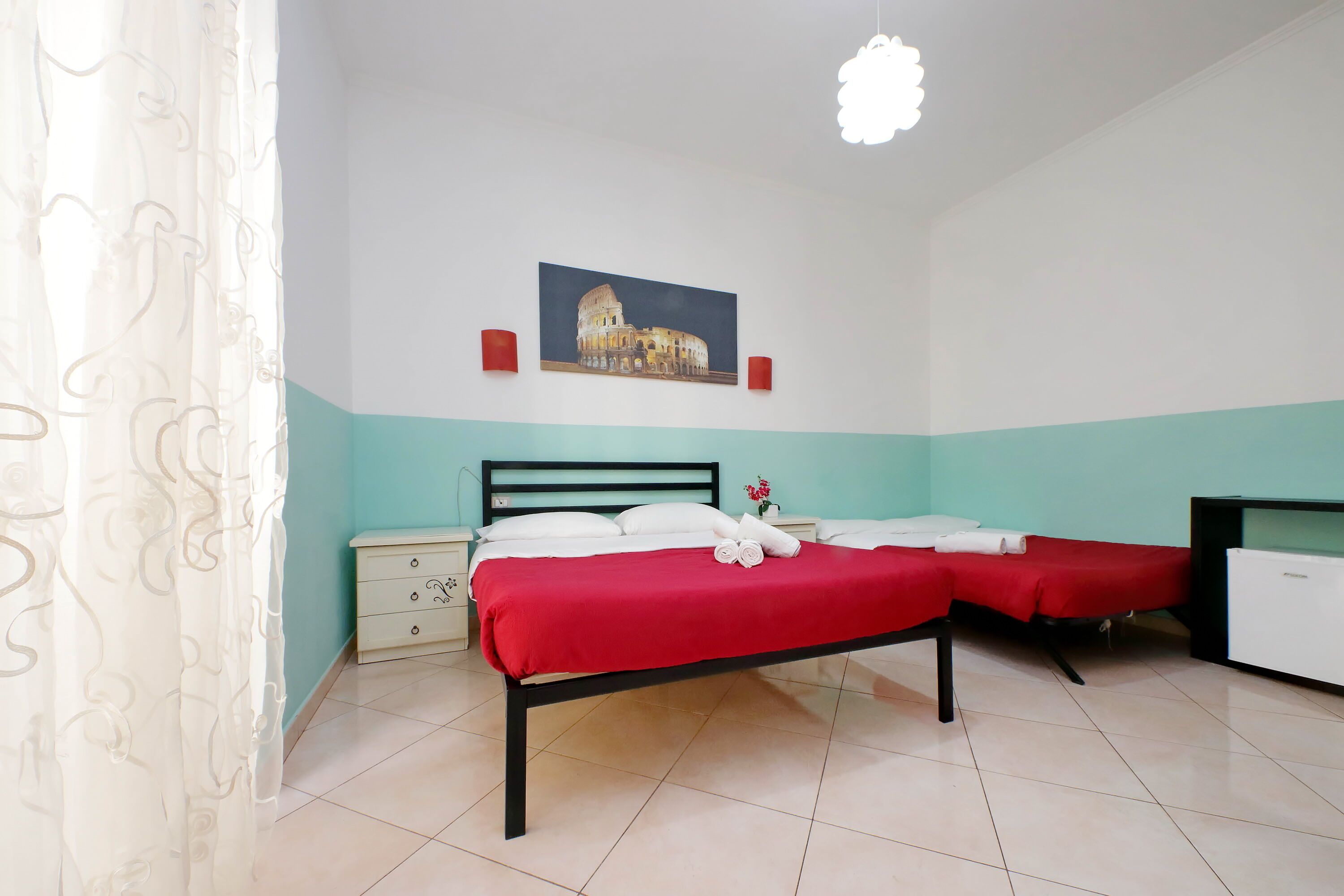 A hotel room with a bed, two bedside tables, a nightstand, a painting of the Colosseum, and a white ceiling light.