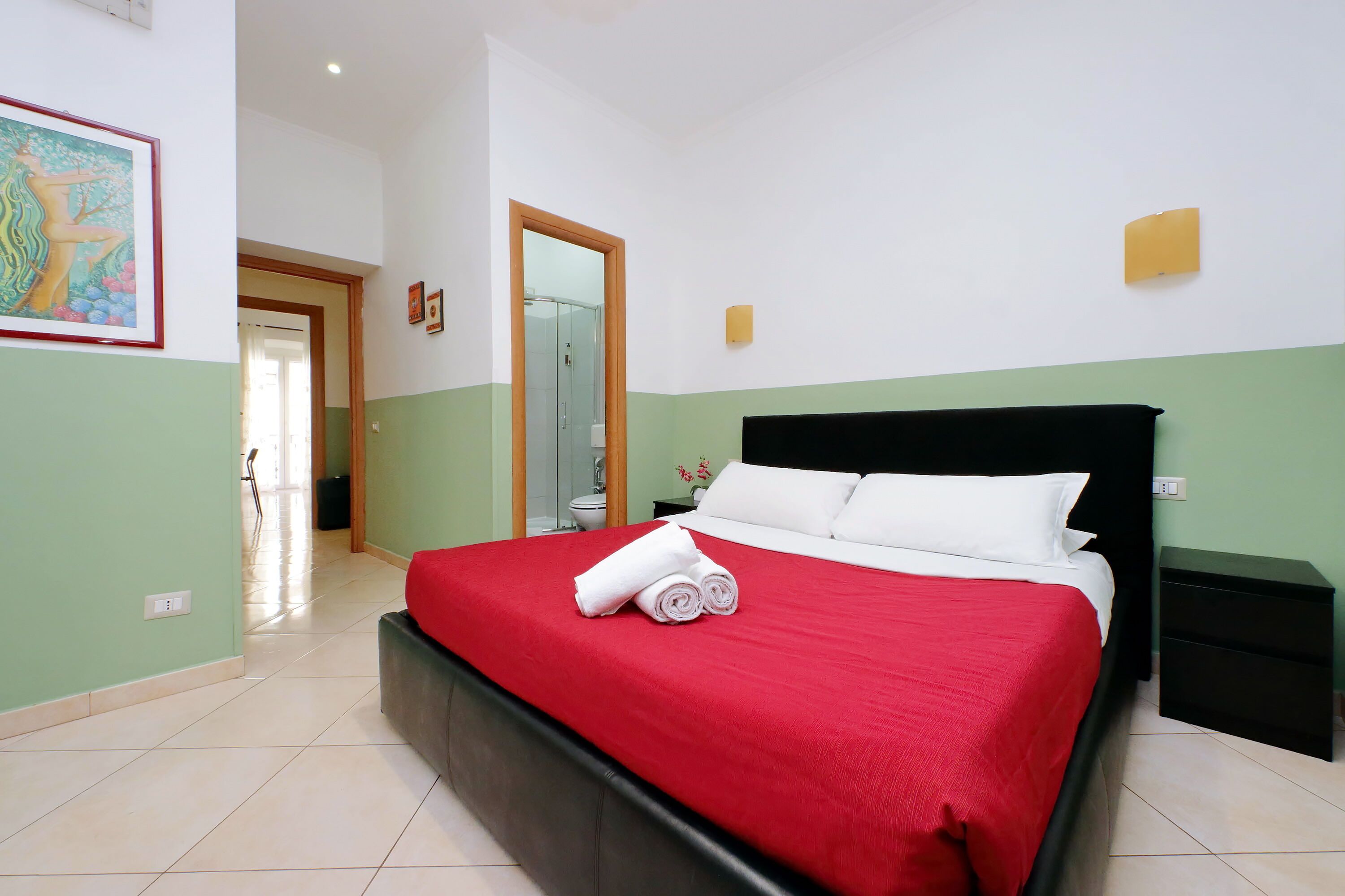 A neatly made bed with a red bedspread and white pillows, a black nightstand, and a green and white wall with a framed picture.