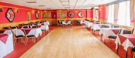 Un amplio salón de banquetes con filas de mesas y sillas, paredes rojas y suelo de madera.