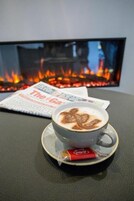 Een kop koffie met latte-art op een tafel, een krant en een open haard op de achtergrond.