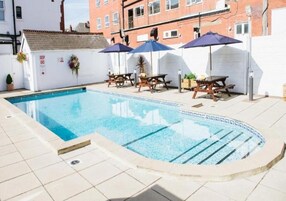 Een zwembad met ligbedden en parasols, omgeven door een witte muur en een bakstenen gebouw op de achtergrond.
