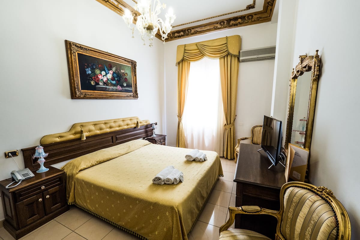 Ein Hotelzimmer mit einem großen Bett, einem Nachttisch mit Telefon, einem Sessel, einem Schreibtisch mit Fernseher und einem Spiegel.