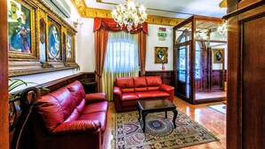 Una habitación con un sofá de cuero rojo, un sillón a juego, una mesa de centro de madera y un tapiz estampado. Se observan obras de arte enmarcadas en las paredes y un candelabro sobre el sofá.