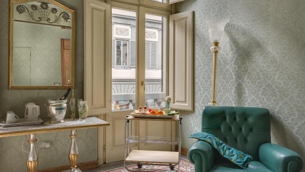 A room with a green armchair, a small table with a tray of food, a mirror, and a window with shutters.