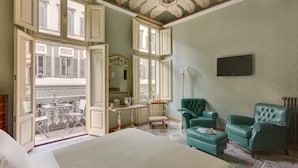 A spacious room with a large bed, two green armchairs, a desk with a chair, a mirror, and a television.