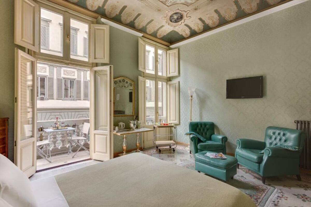 Une pièce spacieuse avec un grand lit, deux fauteuils verts, un bureau avec une chaise, un miroir et un téléviseur.