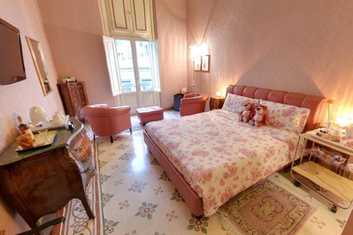 Une chambre à coucher avec une couvre-lit à motifs floraux, une table de chevet en bois avec une lampe, une chaise et une petite table sur laquelle se trouve un plateau avec divers objets.
