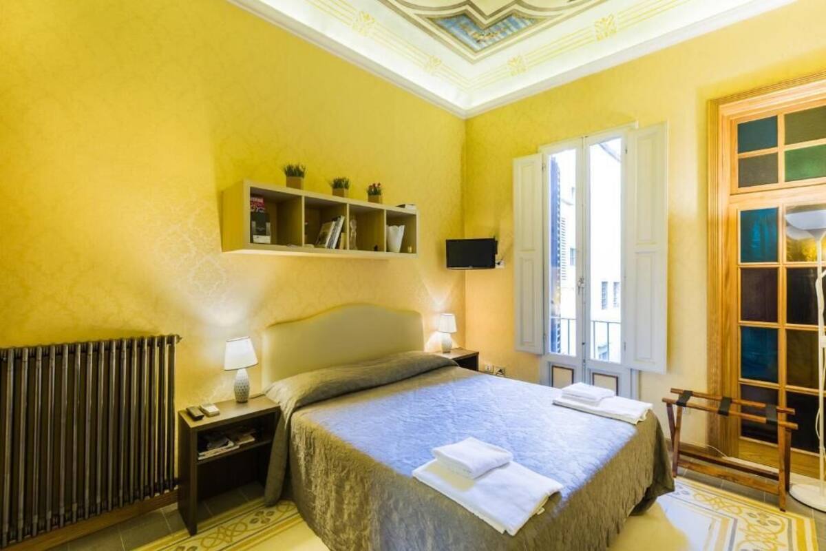 Une chambre d’hôtel avec un lit, des lampes de chevet, une étagère avec des livres et des plantes, une télévision et une fenêtre donnant sur un beau paysage.