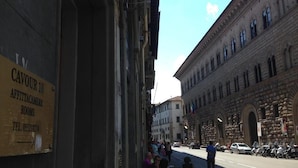 A street view with a building labeled "CAVOUR 10 AFFITTACAMERIO ROOMS" and a street with parked cars and people.