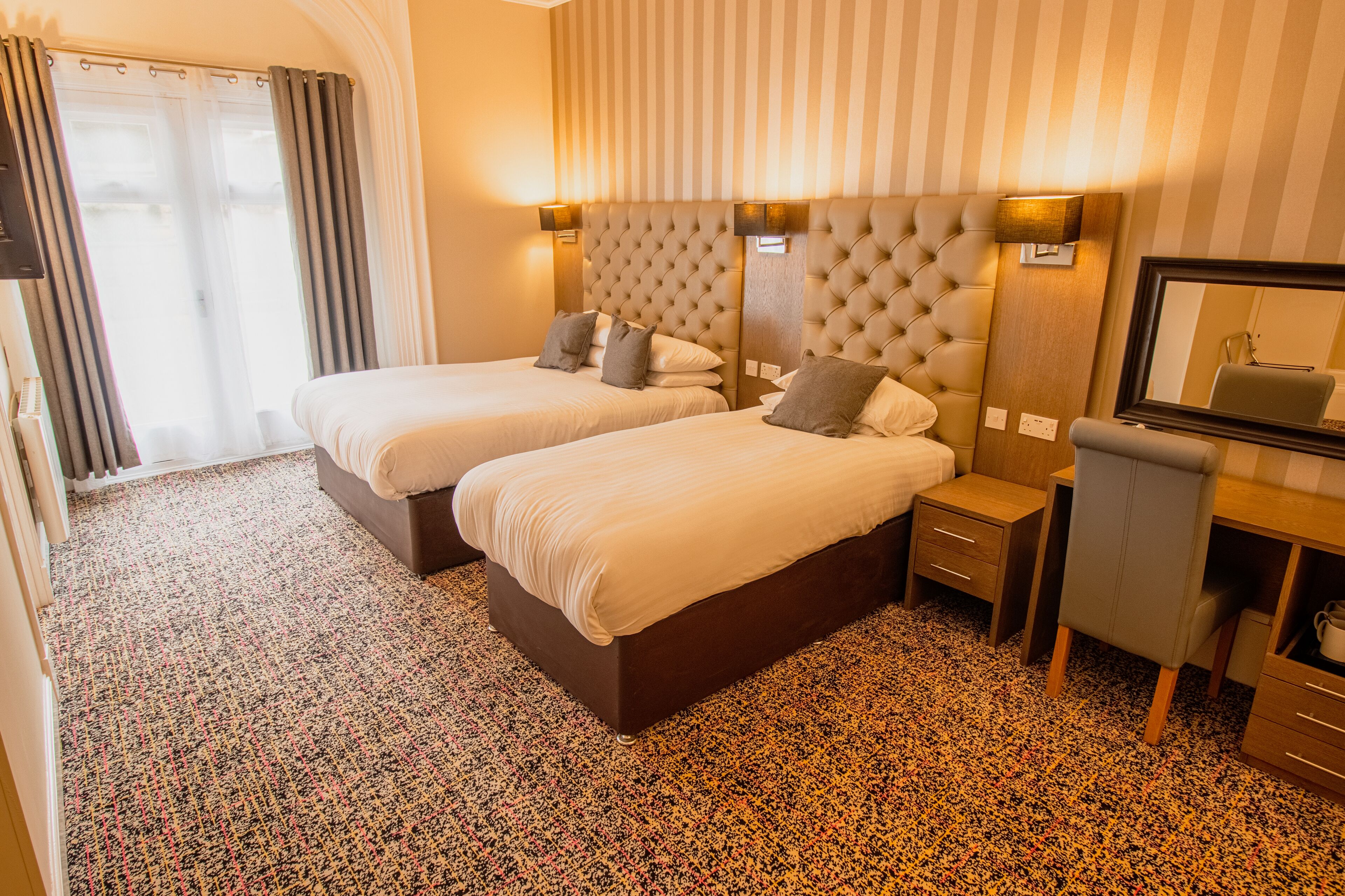 Ein Hotelzimmer mit zwei Betten, einem Schreibtisch, einem Stuhl, einem Spiegel und einem Fenster mit Vorhängen.