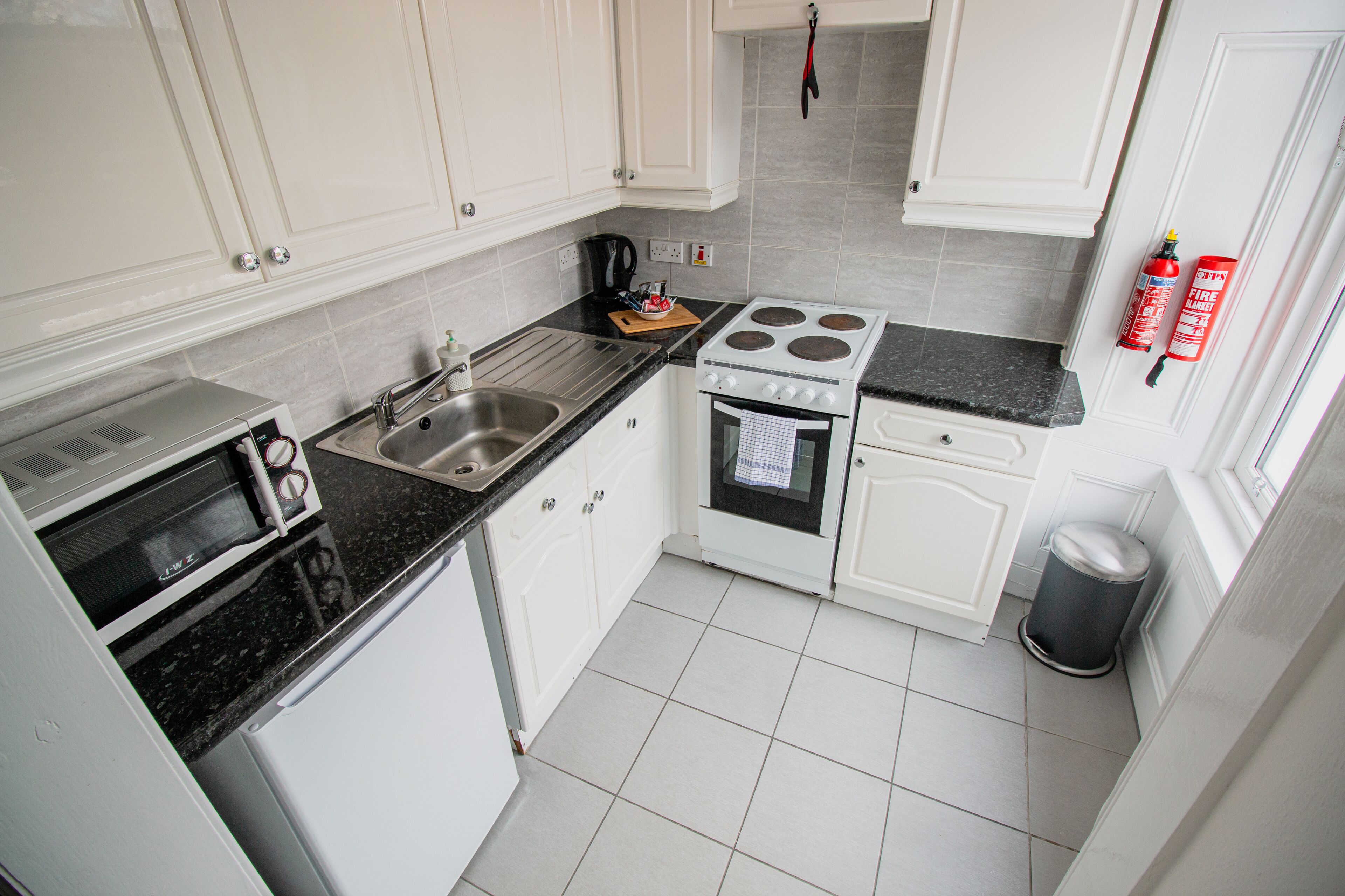 Una cocina compacta con armarios blancos, estufa, fregadero y cubo de basura.