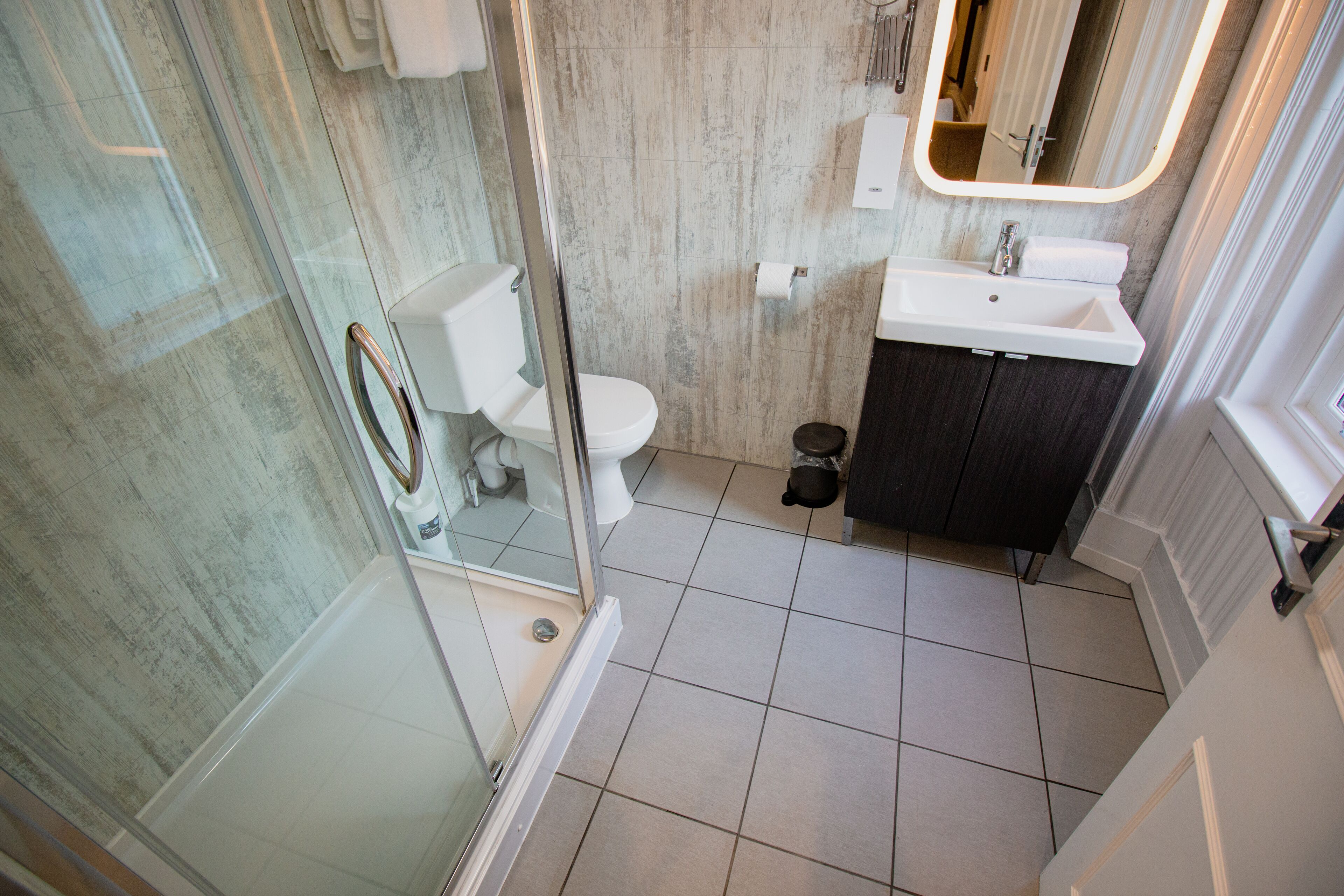 A bathroom with a glass shower enclosure, a white toilet, a sink with a dark cabinet, and a mirror with a lighted frame.