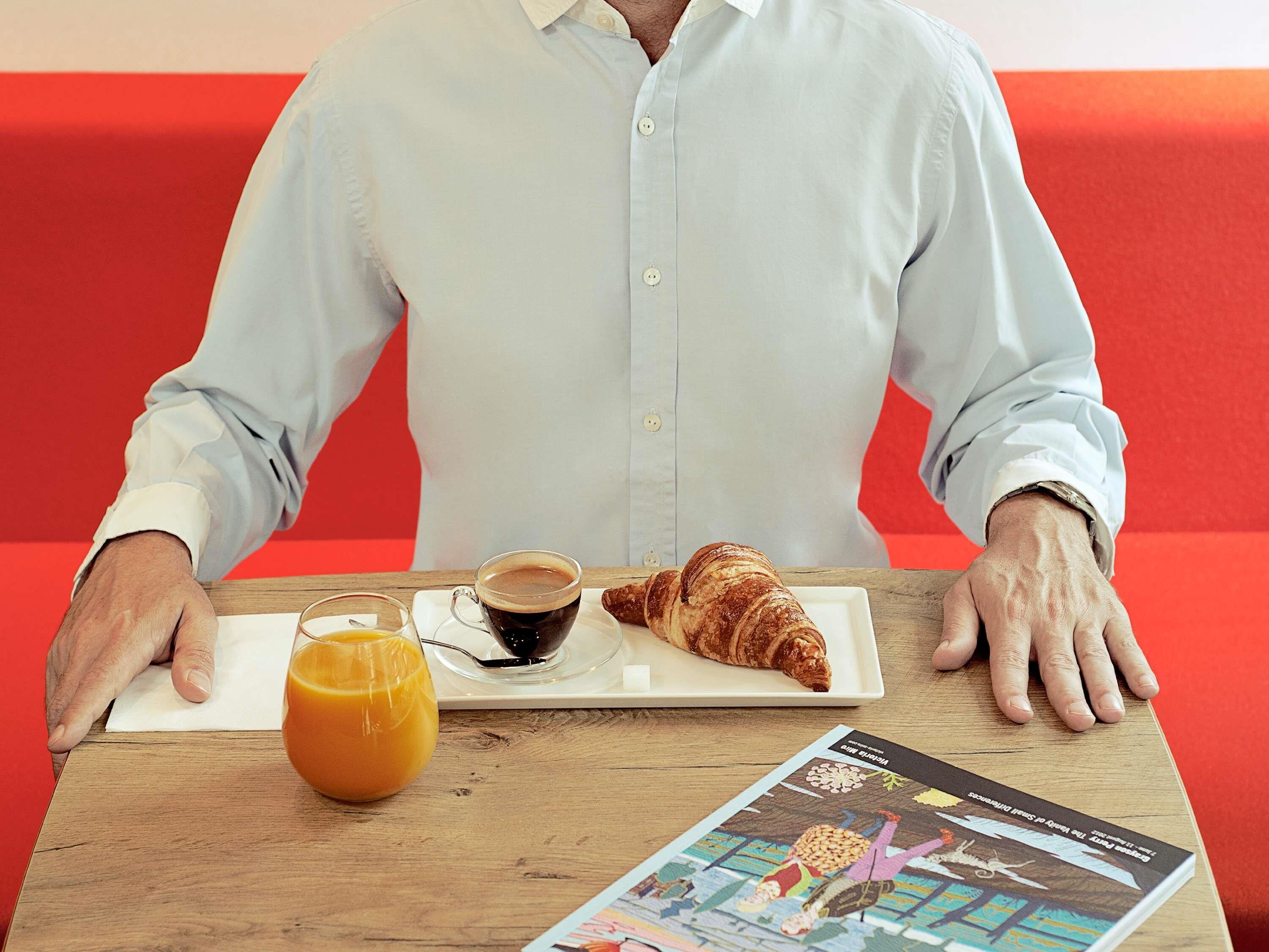 Eine Person sitzt an einem Tisch mit einem Croissant, einer Tasse Kaffee und einem Glas Orangensaft.