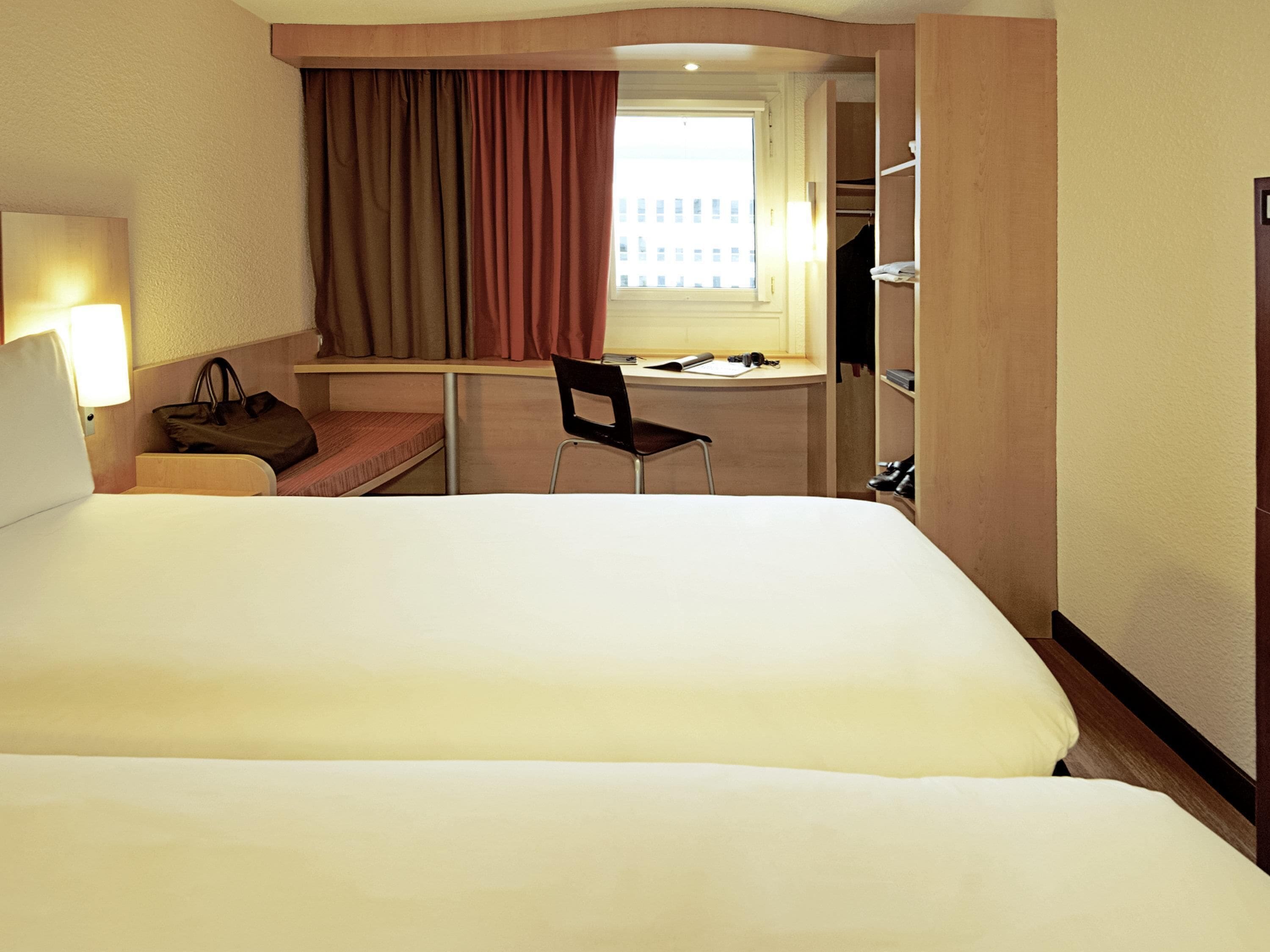 A hotel room with a large bed, a desk, a chair, a desk lamp, a handbag, and a window with curtains.