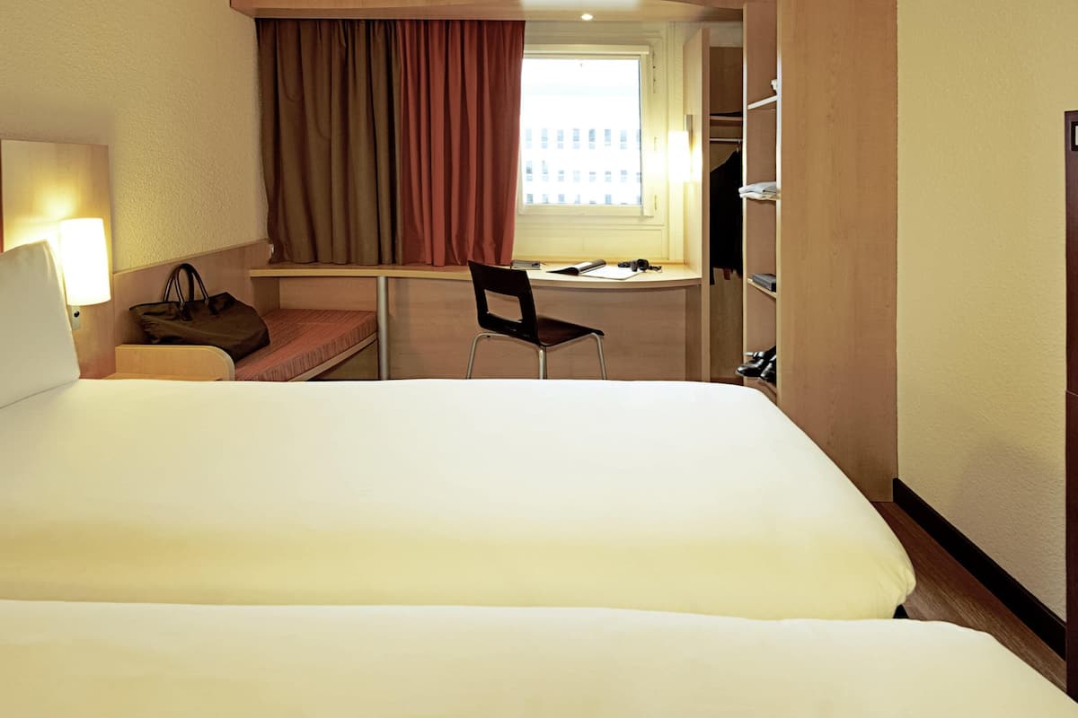 A hotel room with a large bed, a desk, a chair, a desk lamp, a handbag, and a window with curtains.