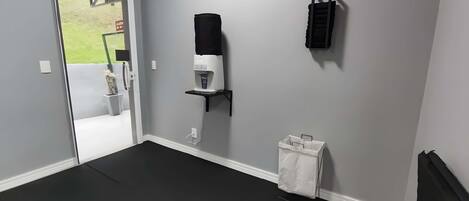 Una habitación con una pared gris, una máquina de café, un estante negro, un cubo de basura blanco y una puerta que da al exterior.