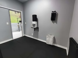 A room with a grey wall, a coffee machine, a black shelf, a white trash bin, and a door leading outside.