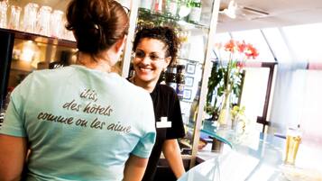 A person wearing a shirt with text, standing in a room with a glass table, and shelves with bottles in the background.