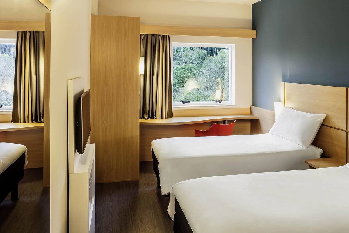 A hotel room with two beds, a desk, a TV, and a window with curtains.
