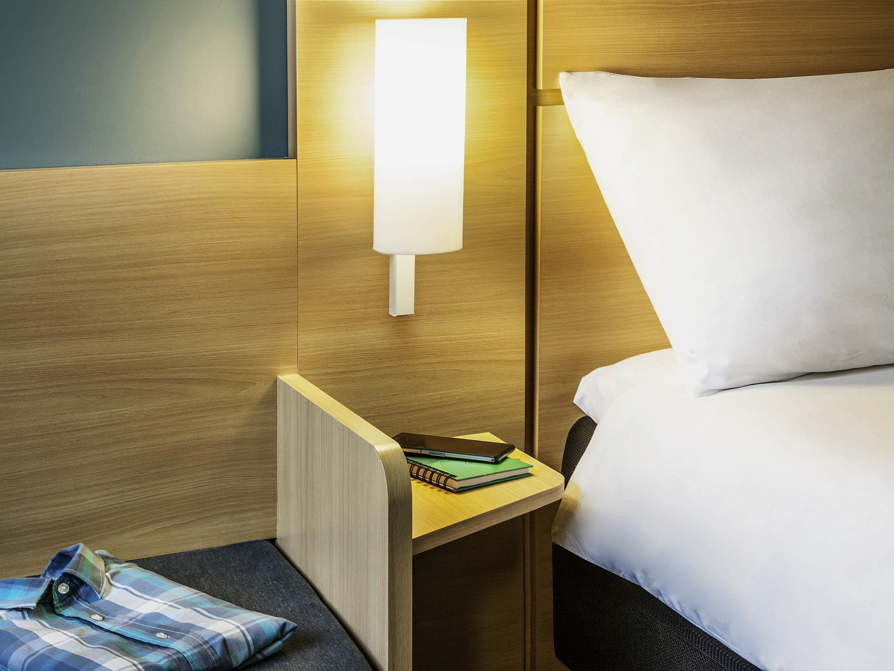A hotel room with a wooden headboard, a bedside table with a lamp, a bed with white bedding, and a small seating area with a pillow and a folded shirt.