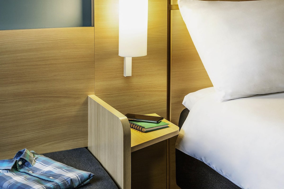 A hotel room with a wooden headboard, a bedside table with a lamp, a bed with white bedding, and a small seating area with a pillow and a folded shirt.