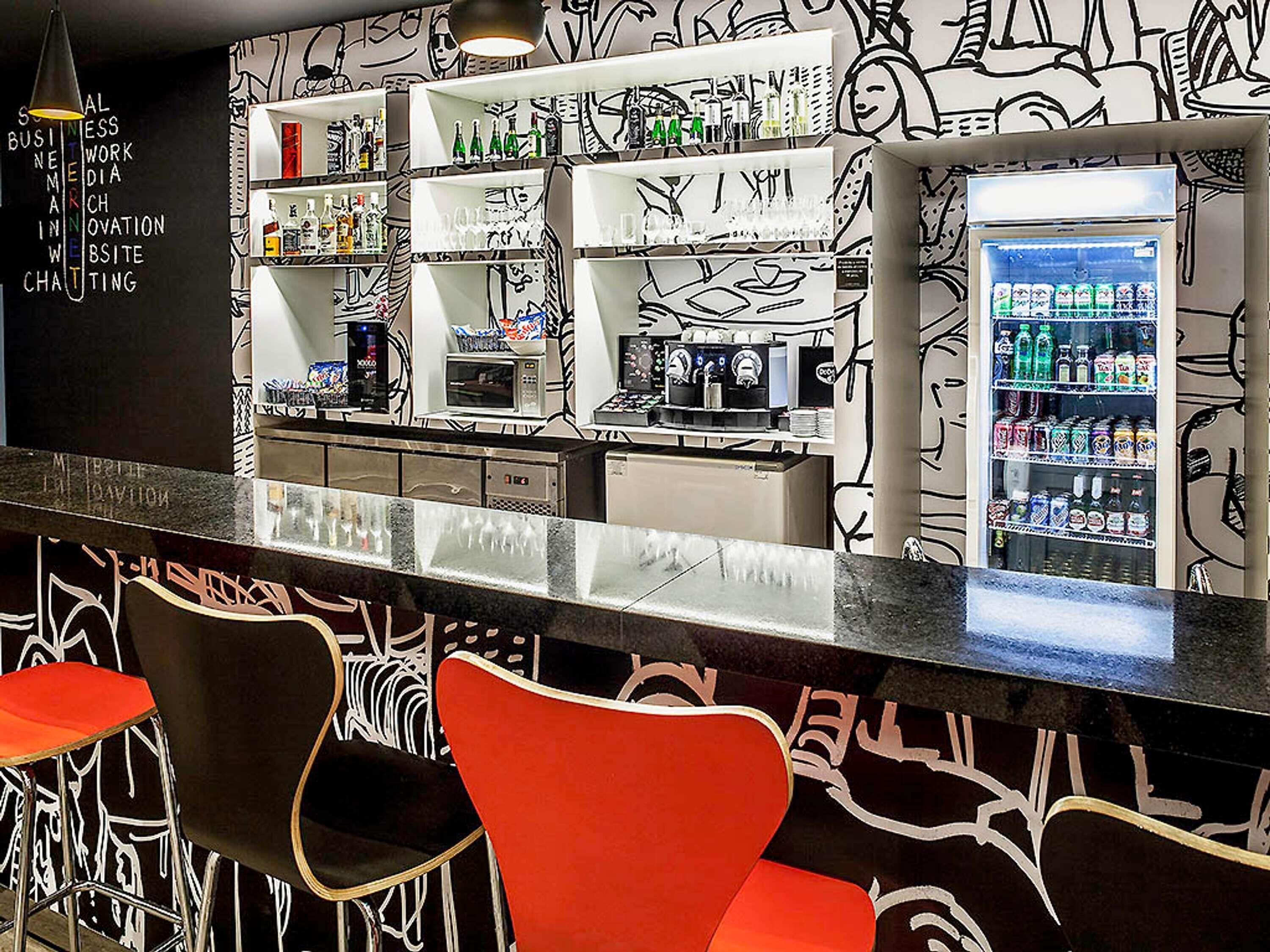 A bar area with a black and white graffiti wall, shelves with bottles, a refrigerator with drinks, and red chairs.