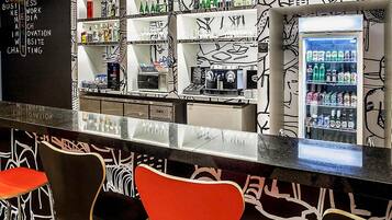 Zona de bar con pared de grafitis en blanco y negro, estanterías con botellas, un refrigerador con bebidas y sillas rojas.