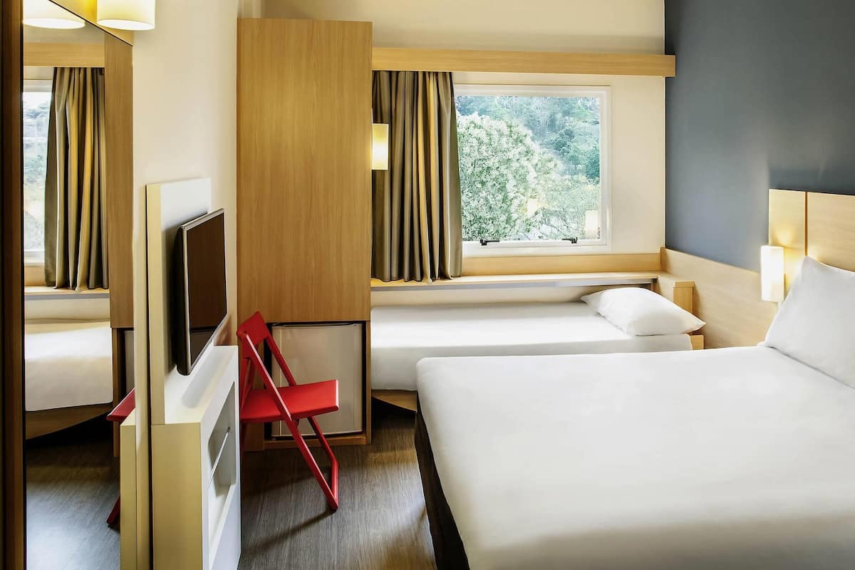A hotel room with a bed, a TV, a red chair, a window with curtains, and a view of trees.