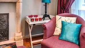 A cozy living room with a fireplace, a red armchair, a small table with a lamp, and a framed artwork on the wall.