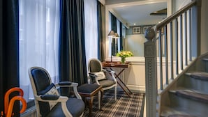 A hotel room with a plaid carpet, two upholstered chairs, a small table with a vase, a lamp, and a staircase with a balustrade.