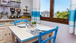 A dining area with a table set for two, a window with curtains, and a kitchen area with food dispensers and a fruit bowl. - Faunele (Orosei)