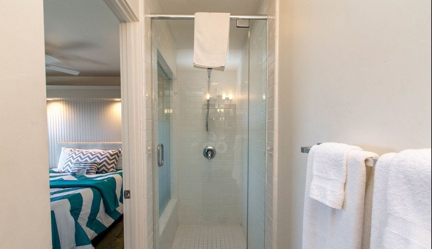Un bany amb una cabina de dutxa de vidre, una barra de tovalloles blanca i un llit amb roba de llit a ratlles a l'habitació propera.