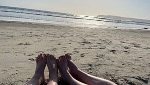 A beach scene with a person's legs and feet resting on the sand, facing the ocean.