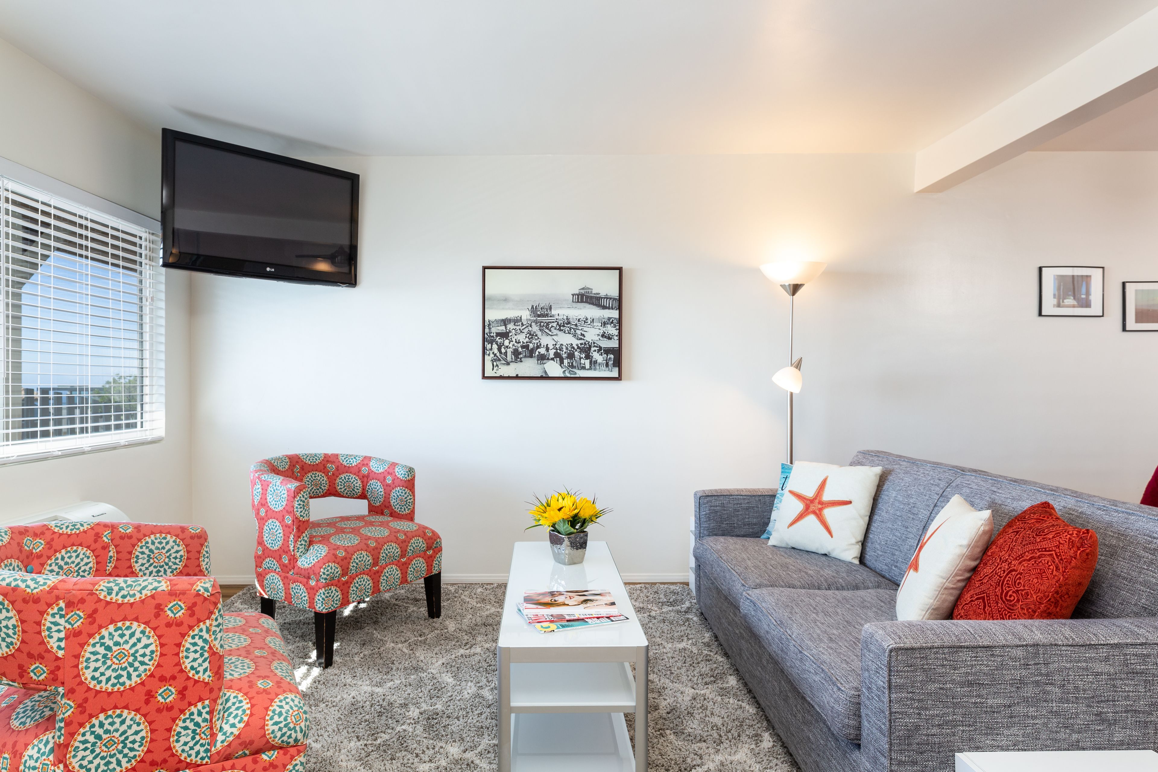 Een woonkamer met een grijze bank, rode fauteuils met een patroon, een witte salontafel met tijdschriften, een zwart lijstje met een foto aan de muur, en een televisie boven een raam met jaloezieën.
