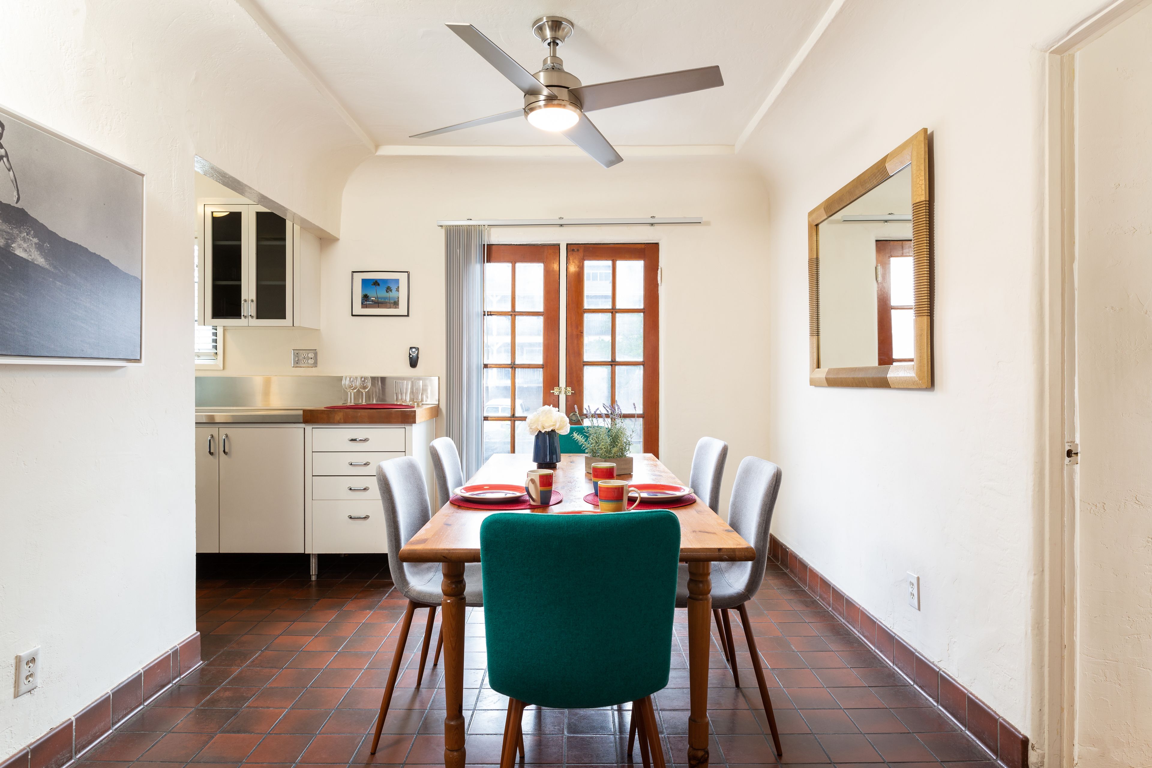 A dining area with a wooden table, chairs, and a ceiling fan. There is a kitchen in the background with cabinets and a window.