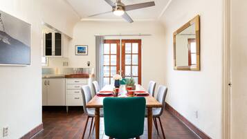 A dining area with a wooden table, chairs, and a ceiling fan. There is a kitchen in the background with cabinets and a window.