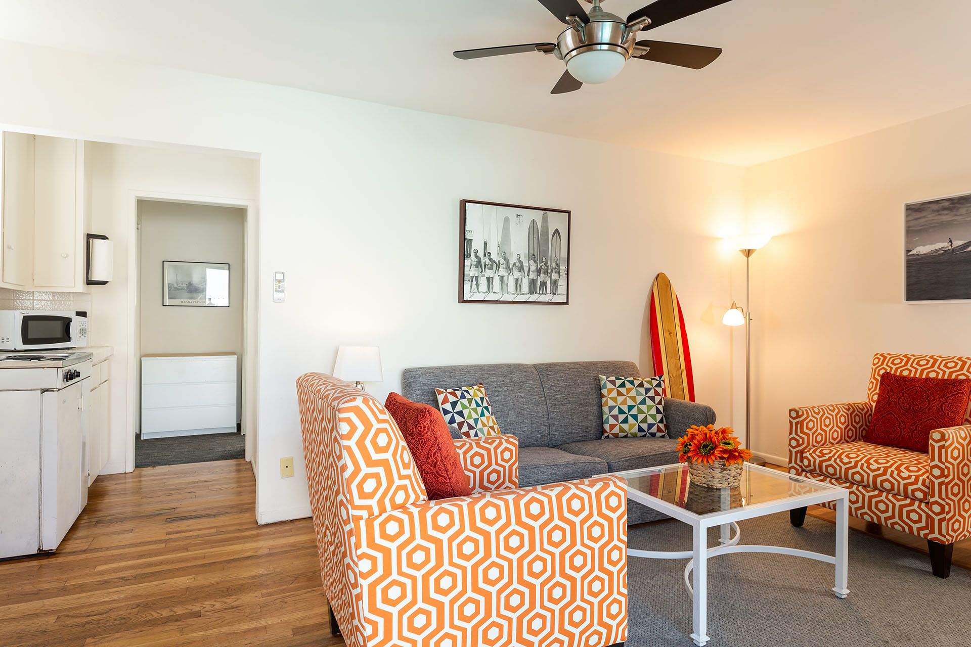 A modern living room with patterned furniture, a ceiling fan, a dining area with a microwave, and a surfboard decoration.