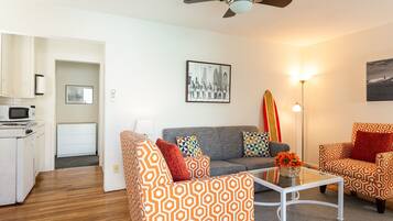 A modern living room with patterned furniture, a ceiling fan, a dining area with a microwave, and a surfboard decoration.