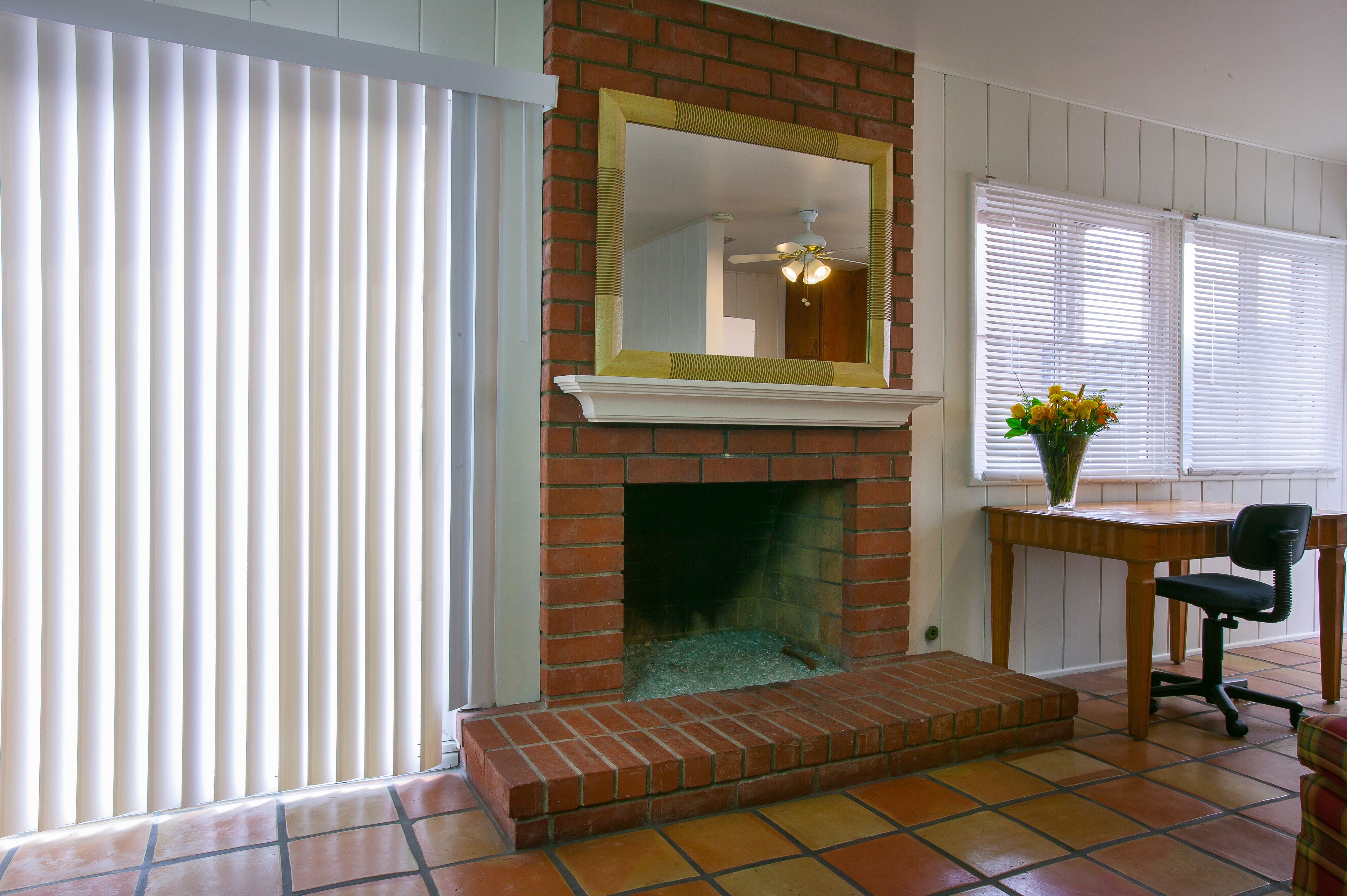 Una habitación con chimenea de ladrillo, un espejo encima, una silla y una ventana con persianas.