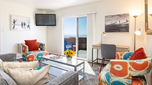 A modern living room with a glass coffee table, colorful patterned sofas, a flat-screen TV, and a view through the glass door.