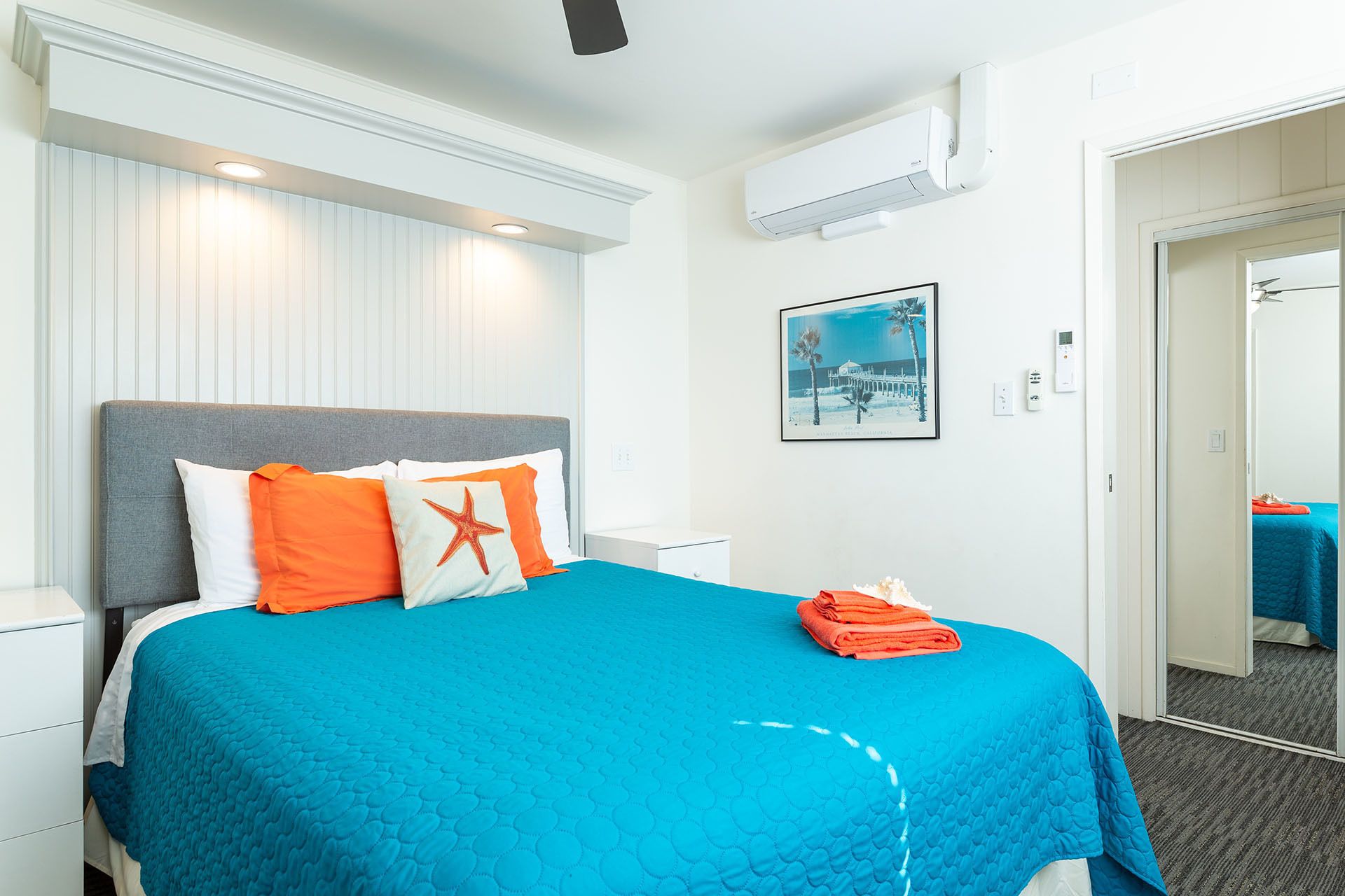 A hotel room with a bed, white nightstands, a blue bedspread, an air conditioning unit, a framed picture, and a door leading to another room.