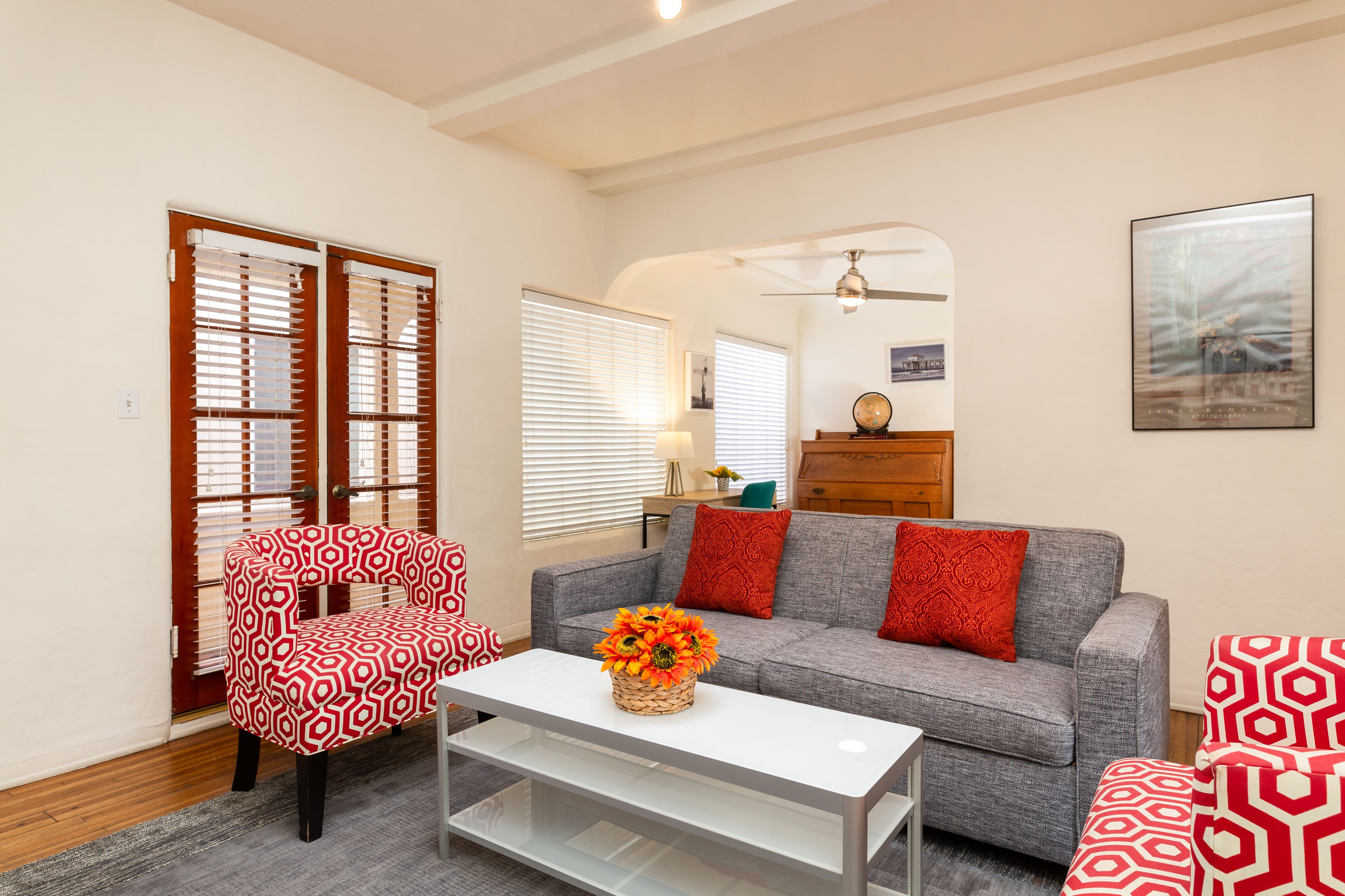 Una sala de estar con un sofá, dos sillones estampados, una mesa de centro con un jarrón de flores y un ventilador de techo.