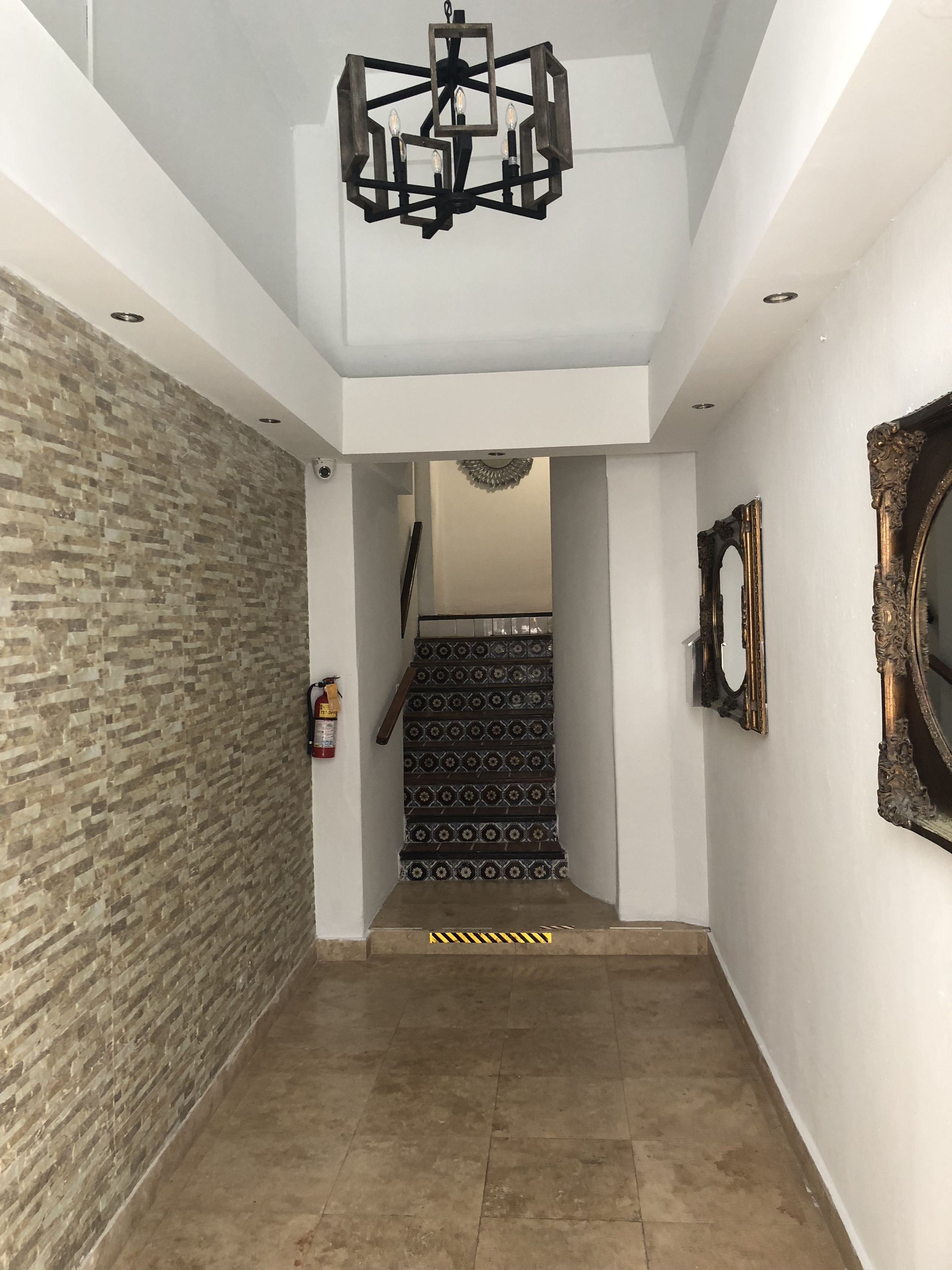 A hallway with a geometric chandelier, patterned stairs, a fire extinguisher, and a mirror on the wall.