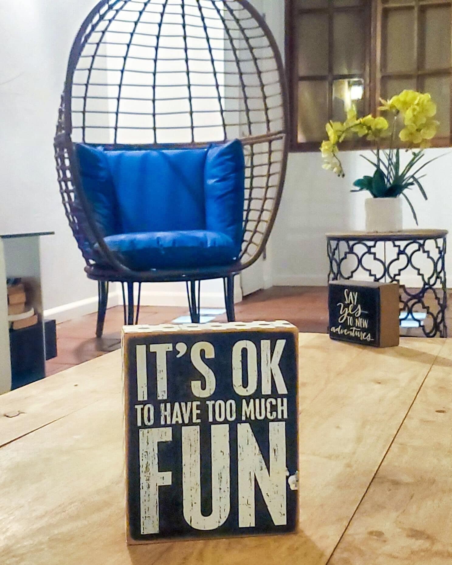 A room with a wooden table, a blue chair, a decorative globe chair, and a sign that reads "IT'S OK TO HAVE TOO MUCH FUN".