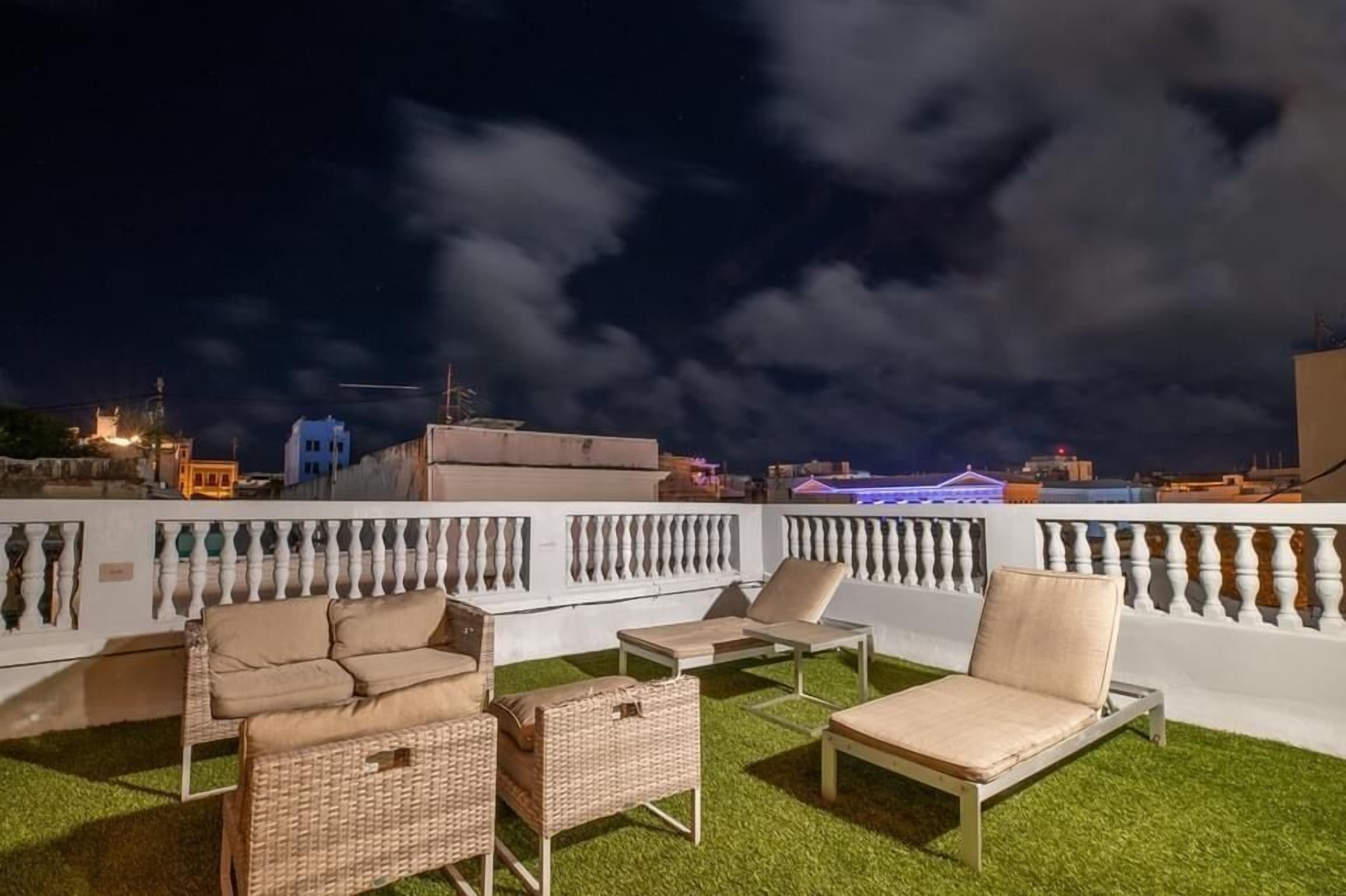 A rooftop terrace with outdoor furniture, a white balustrade, and a cityscape view at night.