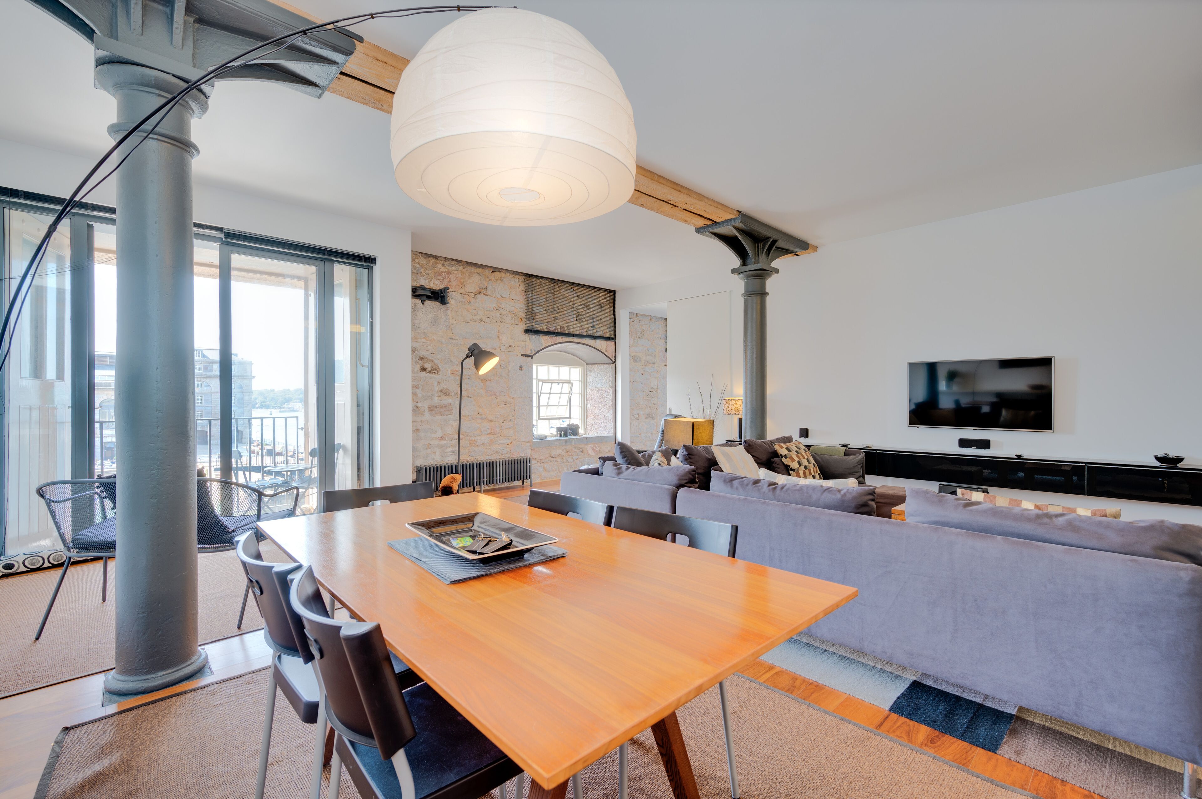A modern living room with a wooden dining table, grey sofa, and a large white pendant light.
