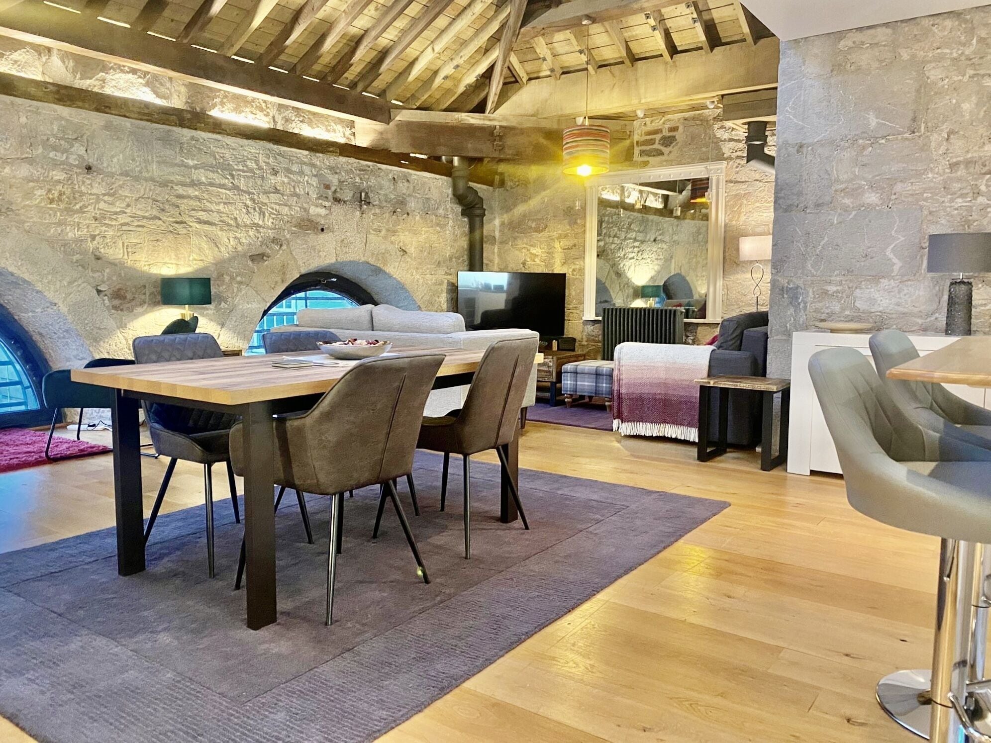 Een rustiek interieur met een stenen muur, houten plafond, een eethoek met stoelen en tafel, een bank en een televisie.